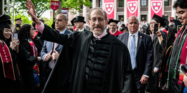 A man in academic robes waves at a graduation ceremony, surrounded by graduates and officials. Harvard banners and a crowd are visible in the background.