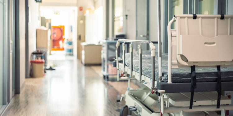 Empty hospital bed with raised side rails positioned in a bright, clean hallway, next to closed doors and medical equipment along the corridor walls.