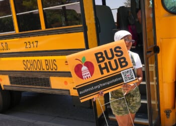 A person stands by an Austin school bus holding a Bus Hub sign with a QR code and web address for Austin ISD transportation.