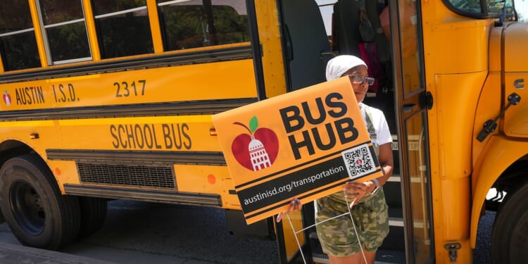 A person stands by an Austin school bus holding a Bus Hub sign with a QR code and web address for Austin ISD transportation.