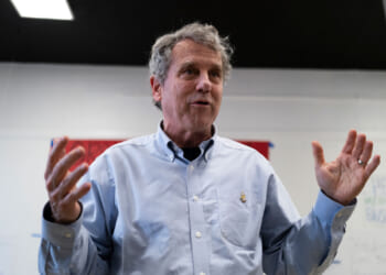 Sherrod Brown with gray hair in a light blue button-down shirt gestures with both hands while speaking indoors, standing in front of a whiteboard and red panel.