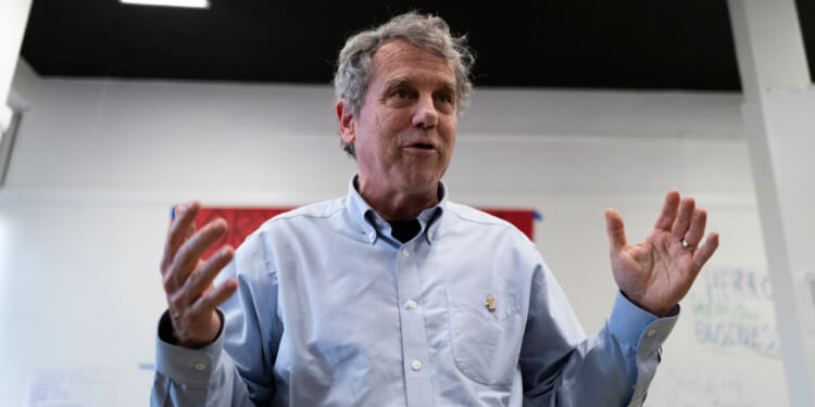 Sherrod Brown with gray hair in a light blue button-down shirt gestures with both hands while speaking indoors, standing in front of a whiteboard and red panel.