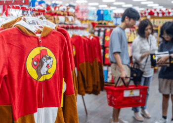 Red and brown animal onesies with a beaver logo hang in a store, while three people with red shopping baskets browse items in the background.