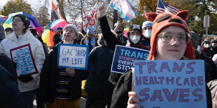 A group of people at a rally hold signs supporting transgender rights, including TRANS HEALTHCARE SAVES LIVES and GAC SAVED MY LIFE. Many wear hats, masks, and warm clothes; rainbow and trans flags are visible.