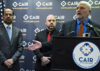 Three men stand at a CAIR podium with microphones. Behind them is a CAIR banner and a U.S. flag. One man is speaking, while another gestures with his hand.