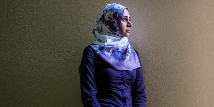 A woman wearing a floral hijab and a dark purple shirt stands against a plain beige wall, looking thoughtfully to the right.