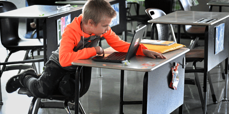 A young boy leans on his desk while using a laptop in a classroom. Papers and supplies are on the desk, and other empty desks are nearby.