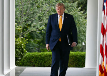 Donald Trump in a dark suit walks along a colonnade holding a folder. Greenery and trees are in the background. The U.S. flag is visible in the foreground on the right.