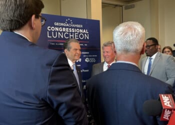 Rick Jackson and Gov. Brian Kemp appear in suits and speak in front of a sign that reads Georgia Chamber Congressional Luncheon while microphones are held toward them for an interview.