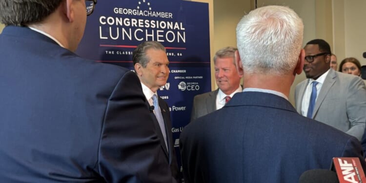 Rick Jackson and Gov. Brian Kemp appear in suits and speak in front of a sign that reads Georgia Chamber Congressional Luncheon while microphones are held toward them for an interview.