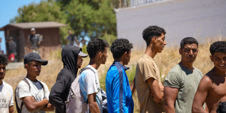 A group of migrants stands in line at El Trampolin beach in Ceuta, Spain, on August 2, 2026, to drink water and fill their bottles from a Spanish Army water truck. People wait to receive drinking water during a humanitarian assistance operation near the coast. (Photo by Ana Lopez/NurPhoto via Getty Images)