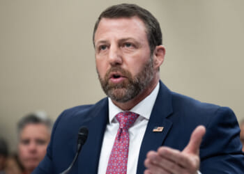 Homeland Security Secretary Markwayne Mullin in a navy suit and pink tie speaks into a microphone at a formal event, gesturing with one hand. He has short brown hair, a beard, and wears an American flag pin on his lapel.