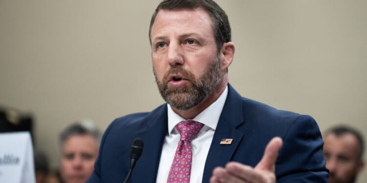 Homeland Security Secretary Markwayne Mullin in a navy suit and pink tie speaks into a microphone at a formal event, gesturing with one hand. He has short brown hair, a beard, and wears an American flag pin on his lapel.