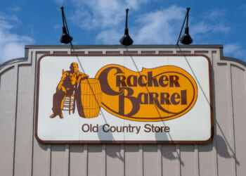 A Cracker Barrel Old Country Store sign on the exterior of a building, featuring a man sitting beside a barrel, with the sky and clouds in the background.