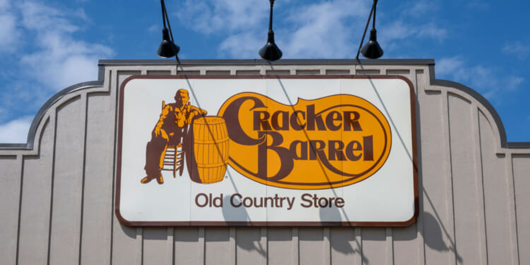 A Cracker Barrel Old Country Store sign on the exterior of a building, featuring a man sitting beside a barrel, with the sky and clouds in the background.