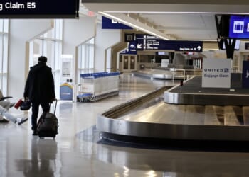 An airport baggage claim area with empty carousels, a few people with luggage, and signs for United and JetBlue. Rows of luggage carts are lined up near seating. Overhead signs display Bag Claim E5..