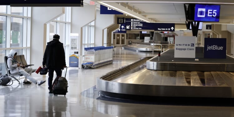 An airport baggage claim area with empty carousels, a few people with luggage, and signs for United and JetBlue. Rows of luggage carts are lined up near seating. Overhead signs display Bag Claim E5..