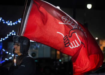 A person in a fur hat holds a lit candle and a large red flag with a white hand design, standing outdoors at night with blue lights blurred in the background.