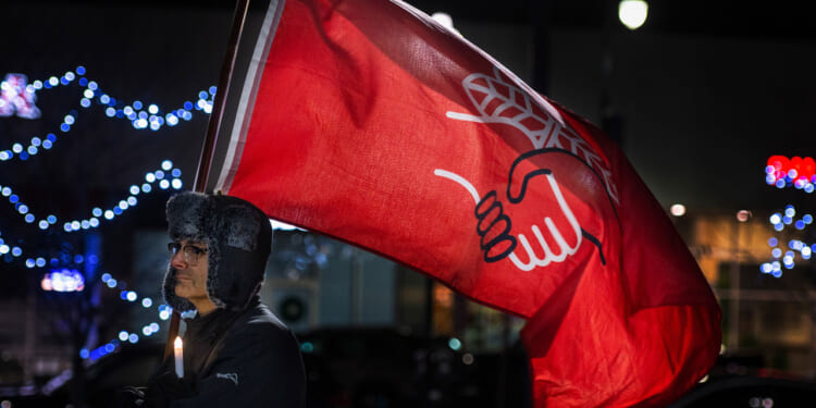 A person in a fur hat holds a lit candle and a large red flag with a white hand design, standing outdoors at night with blue lights blurred in the background.