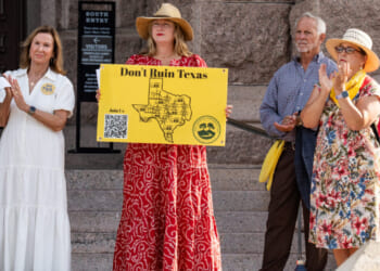 Four adults stand on stone steps at a rally. One woman in a red dress and hat holds a yellow sign reading “Don’t Ruin Texas,” protesting concerns over the new Data Center project. The others wear summer clothes, and a stone building is visible in the background.