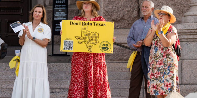 Four adults stand on stone steps at a rally. One woman in a red dress and hat holds a yellow sign reading “Don’t Ruin Texas,” protesting concerns over the new Data Center project. The others wear summer clothes, and a stone building is visible in the background.