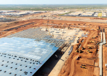 Aerial view of a large data center construction site with a partially completed building, cranes, machinery, and piles of materials on reddish-brown soil, surrounded by other industrial facilities.