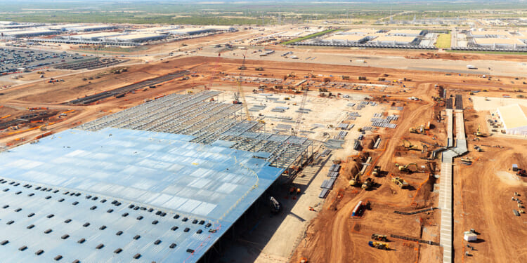 Aerial view of a large data center construction site with a partially completed building, cranes, machinery, and piles of materials on reddish-brown soil, surrounded by other industrial facilities.