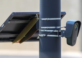 A solar panel and a surveillance camera are mounted on a black pole using metal straps, with a beige wall blurred in the background.