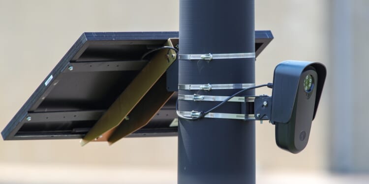 A solar panel and a surveillance camera are mounted on a black pole using metal straps, with a beige wall blurred in the background.