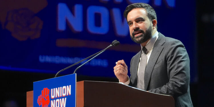 Democratic Socialists of America member and New York City Mayor Zohran Mamdani speaks behind a "union now" poster. Pictured: A man in a suit speaks at a podium with a Union Now sign, in front of a large Union Now banner.