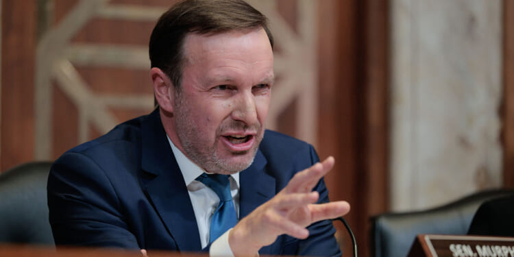 Sen. Chris Murphy's plan to increase the minimum wage would harm low-income workers, a study finds. Pictured: Murphy, a man in a blue suit speaks emphatically at a desk with a nameplate reading SEN. MUR visible, gesturing with his hand. The background shows a wooden panel and part of a marble wall.