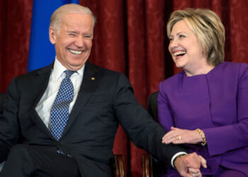 Two people in business attire sit side by side, smiling and laughing. The person on the left wears a dark suit and patterned tie; the person on the right wears a purple outfit. They are holding hands and appear to be enjoying a light moment.