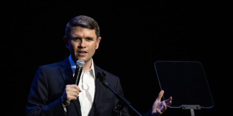 James Talarico in a suit speaks into a microphone on stage, gesturing with one hand, with a transparent teleprompter next to him against a dark background.