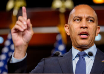 A man in a suit and tie speaks, raising his index finger. Blurred U.S. flags and a dark background are visible behind him.