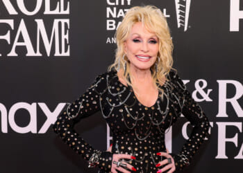 Dolly Parton with long blonde hair in a studded black outfit smiles on a red carpet, posing in front of a backdrop with Rock & Roll Hall of Fame and sponsor logos.