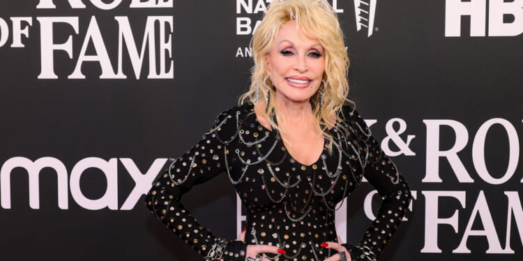 Dolly Parton with long blonde hair in a studded black outfit smiles on a red carpet, posing in front of a backdrop with Rock & Roll Hall of Fame and sponsor logos.