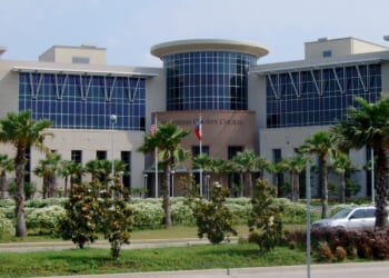 A modern courthouse building with large windows, surrounded by palm trees and greenery, with a few cars parked nearby and American and Texas flags at the entrance.