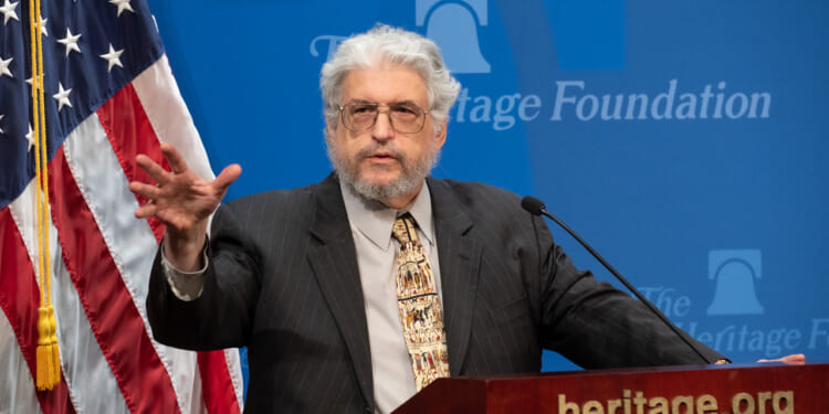 An older man with gray hair and a beard speaks at a podium with a microphone, gesturing with one hand. An American flag and a Heritage Foundation logo are visible in the background.