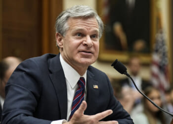 A new report sheds more light on the Richmond Catholic Memo. Pictured: FBI Director Christopher Wray, a man in a suit speaks into a microphone at a hearing, gesturing with one hand. People and a U.S. flag are visible in the background, along with a large framed portrait.