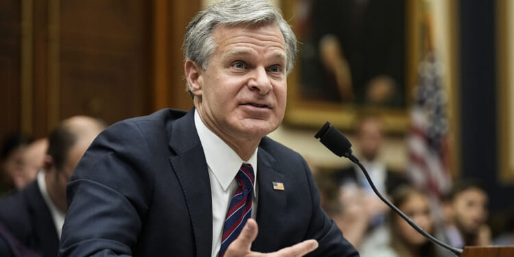 A new report sheds more light on the Richmond Catholic Memo. Pictured: FBI Director Christopher Wray, a man in a suit speaks into a microphone at a hearing, gesturing with one hand. People and a U.S. flag are visible in the background, along with a large framed portrait.