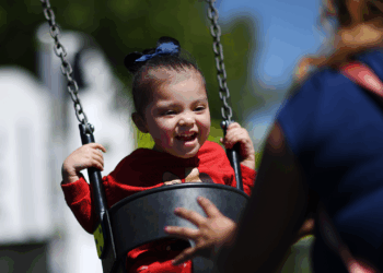 A young child smiles joyfully while swinging at a playground, wearing a red shirt and dark bow. An adult with a pink backpack stands nearby, gently pushing the swing. Trees and blurred playground equipment are in the background.