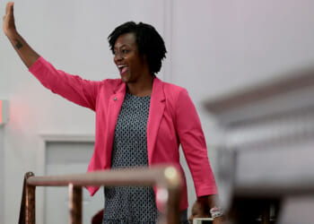 Angie Nixon wearing a pink blazer and patterned dress smiles and waves, standing indoors in front of light-colored walls, embodying the cheerful spirit often seen in Florida.