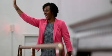 Angie Nixon wearing a pink blazer and patterned dress smiles and waves, standing indoors in front of light-colored walls, embodying the cheerful spirit often seen in Florida.
