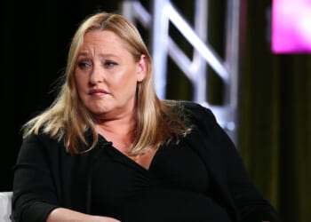 Former SPLC CFO Heidi Beirich was arrested Tuesday. Pictured: Beirich, a woman with long blonde hair wearing a black outfit sits in a chair, looking slightly to the side with a serious expression. A large metal truss and colorful lights are in the background.