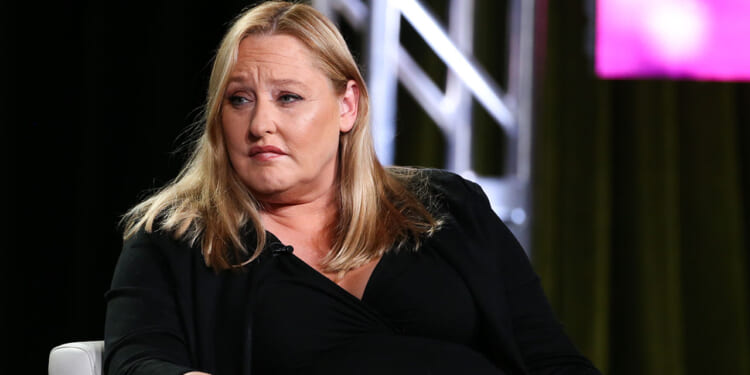 Former SPLC CFO Heidi Beirich was arrested Tuesday. Pictured: Beirich, a woman with long blonde hair wearing a black outfit sits in a chair, looking slightly to the side with a serious expression. A large metal truss and colorful lights are in the background.