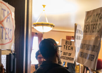 People holding protest signs against data centers indoors, with one sign featuring a large red slash through the words DATA CENTERS and others mentioning BIG TECH and demanding to keep every new data center out of Wisconsin.