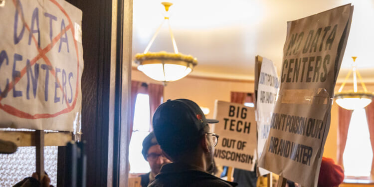 People holding protest signs against data centers indoors, with one sign featuring a large red slash through the words DATA CENTERS and others mentioning BIG TECH and demanding to keep every new data center out of Wisconsin.