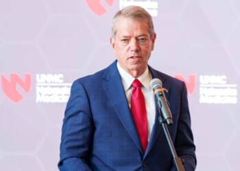 Nebraska Gov. Jim Pillen in a blue suit and red tie speaks into a microphone at a podium, with a blurred backdrop featuring the UNMC Nebraska Medicine logo.