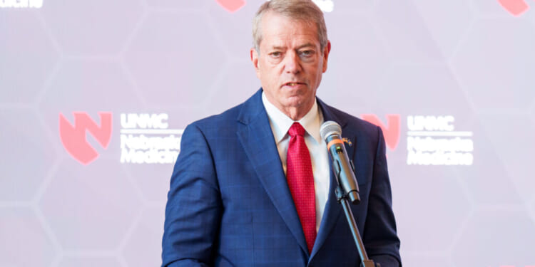 Nebraska Gov. Jim Pillen in a blue suit and red tie speaks into a microphone at a podium, with a blurred backdrop featuring the UNMC Nebraska Medicine logo.