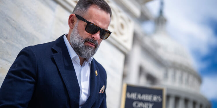 Cory Mills with a beard and sunglasses stands outside a marble building, wearing a dark suit and white shirt. A Members Only sign and a domed structure are visible in the background.